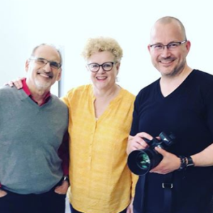 Photo of two men and one woman smiling. One man holds a camera.
