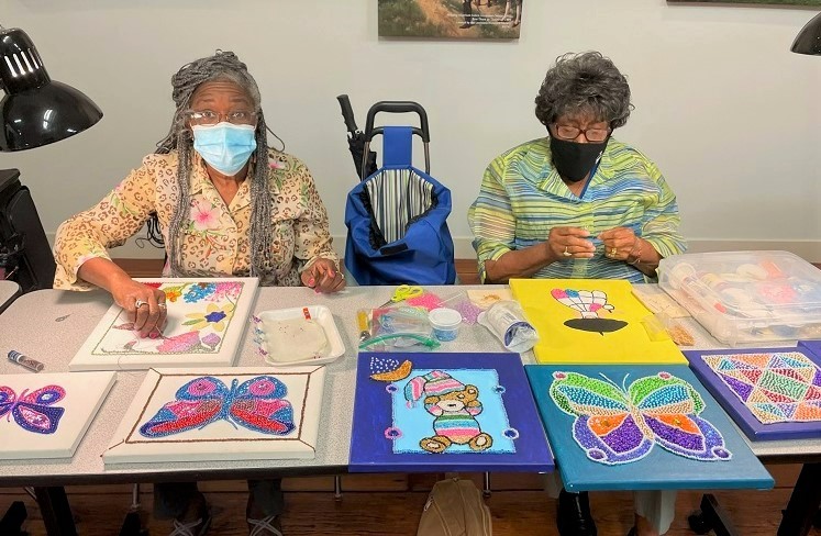 Two older adult participants work on a beading project at a table.